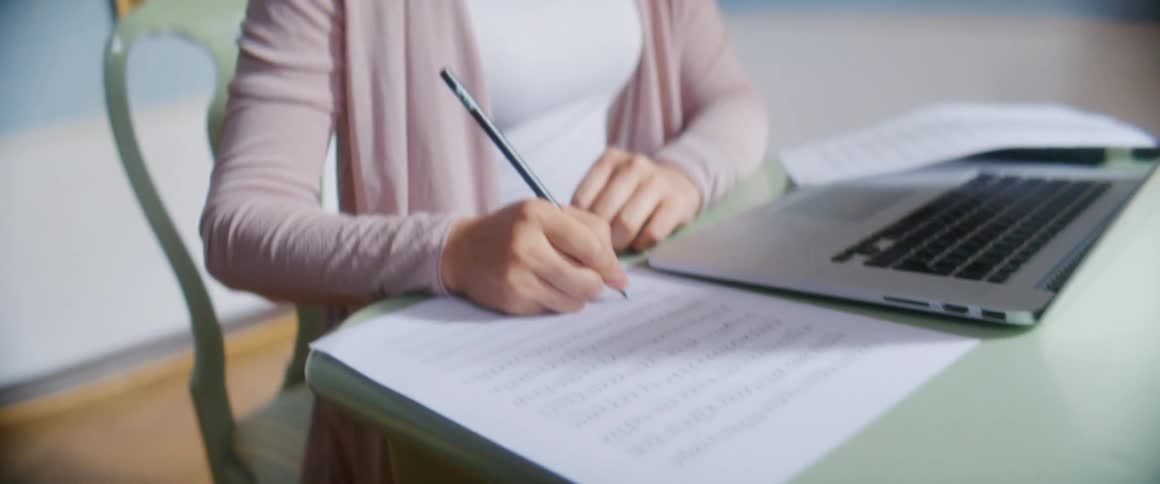 A woman sits at a table with a laptop and writes on a piece of paper