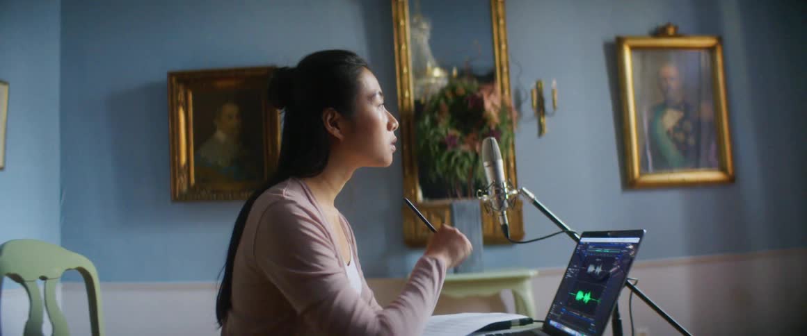 A young woman at home sits at a desk in front of a laptop and writes on a paper and thinks