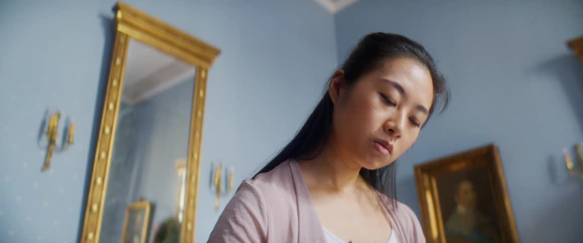 A woman sitting indoors thinks for a moment before writing something with a pen