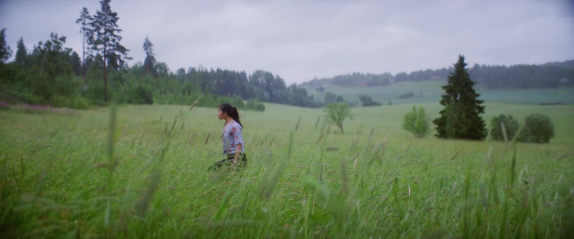 Woman walks through tall grass with forest covered hills nearby and on the horizon