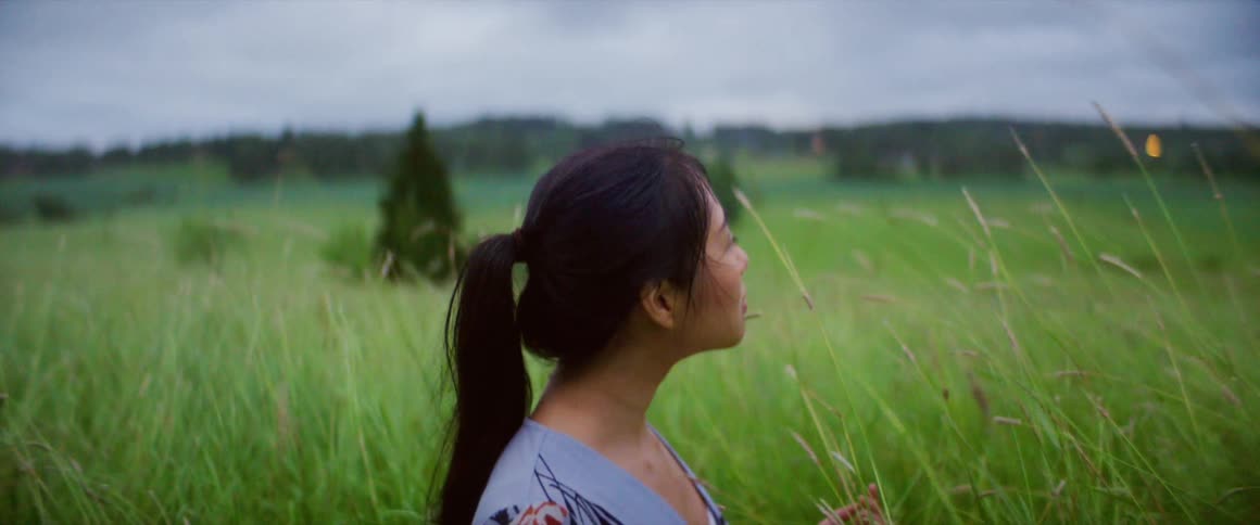 A young asian woman sits among the tall grass in a field on a cloudy day