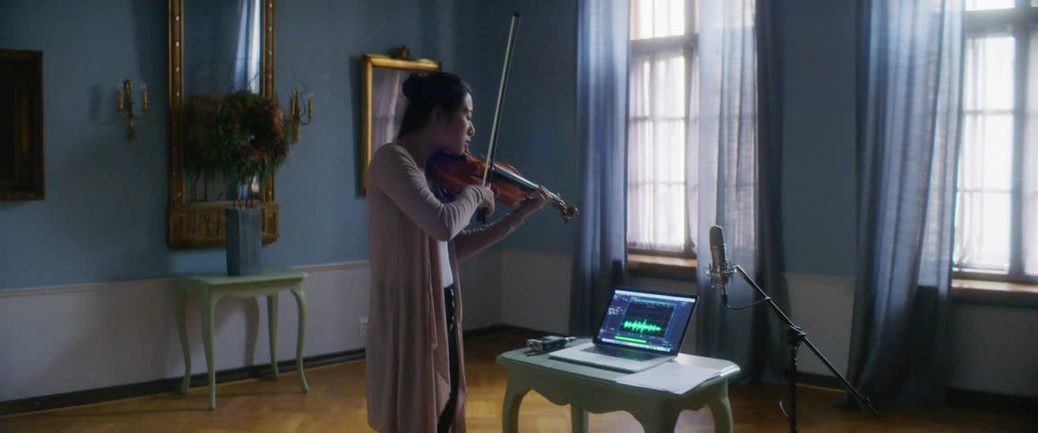 A woman stands in front of a laptop and microphone playing the violin