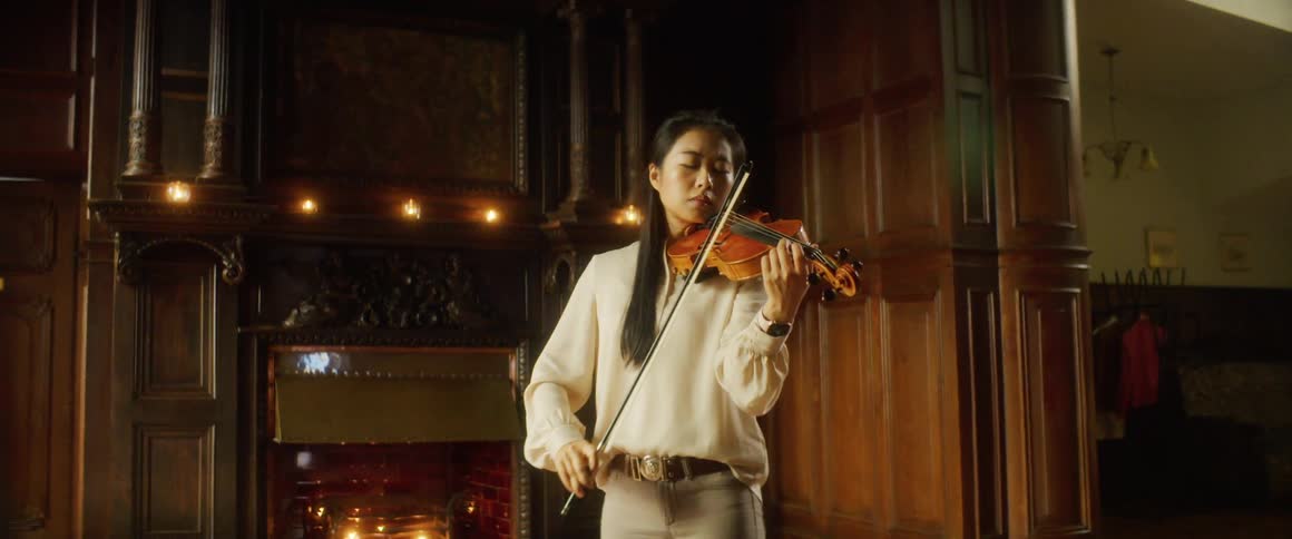 Asian woman holding a violin against her neck and moving a bow plays a violin in front of the fireplace in a room
