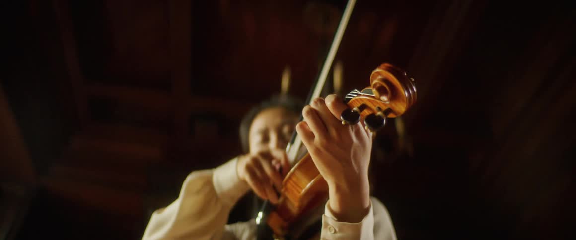 Asian woman holding a violin against her neck and moving a bow plays a violin under a chandelier in a large room