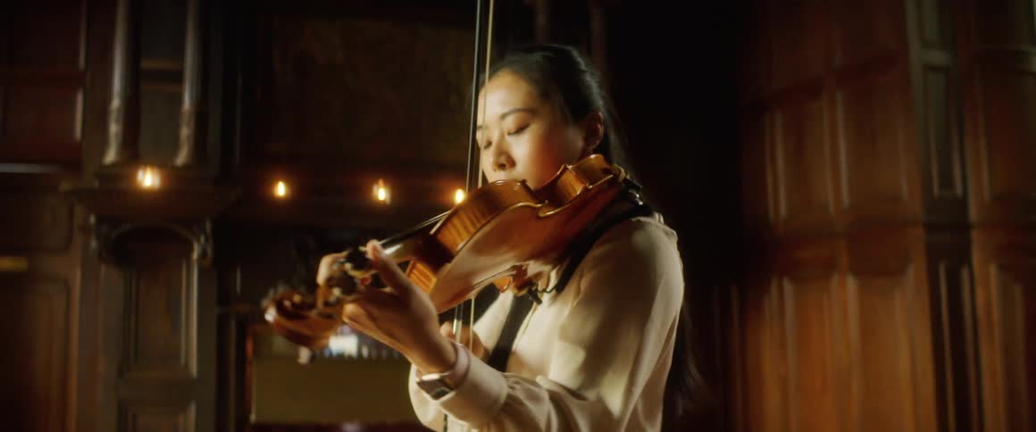 A young woman plays the violin while standing indoors in a building with stained glass windows