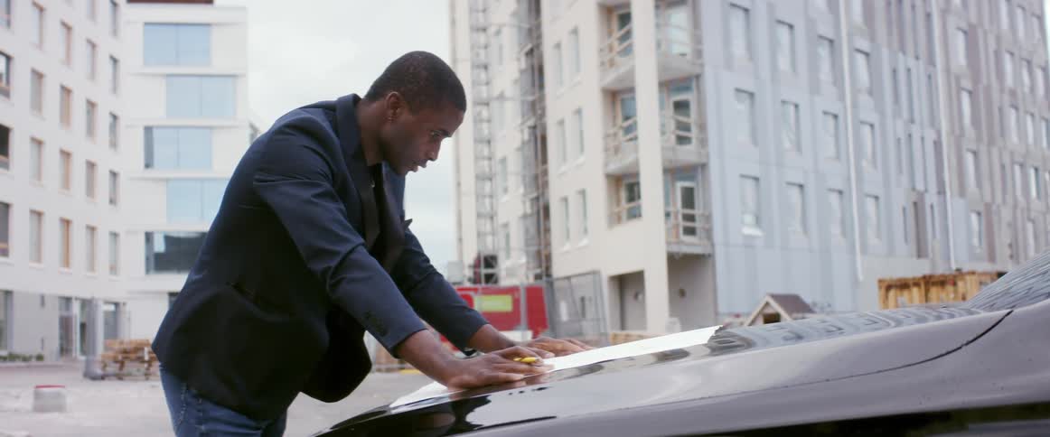 Man reads a blueprint over the hood of a car in a construction site