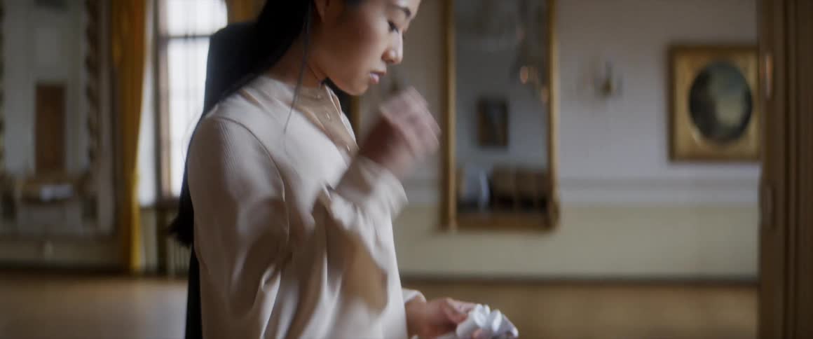 A young woman carrying an instrument case on her back places a pair of wireless ear buds in her ears while standing indoors in a large room during the day