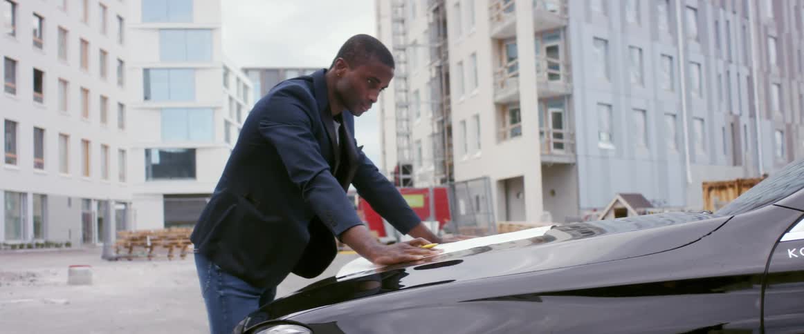 A man leans against a car with a large piece of paper outside of large city buildings