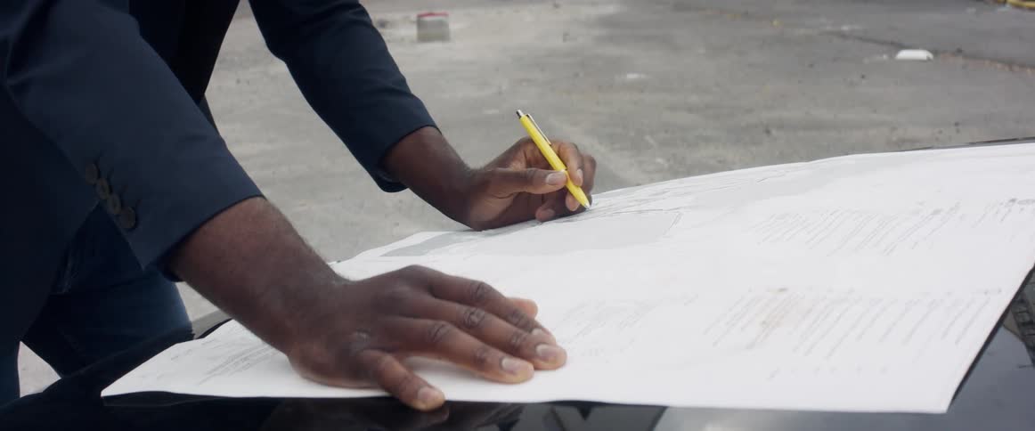 A black person stands outside poised with a pen to write on a piece of paper laying on a flat surface outside