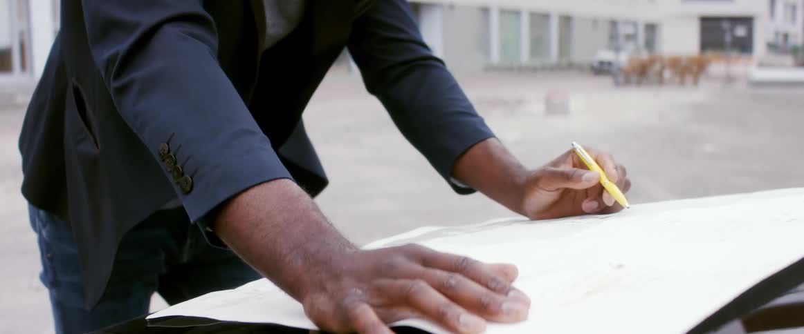 Man in business suit leans over car hood examining paper while holding writing utensil near buildings