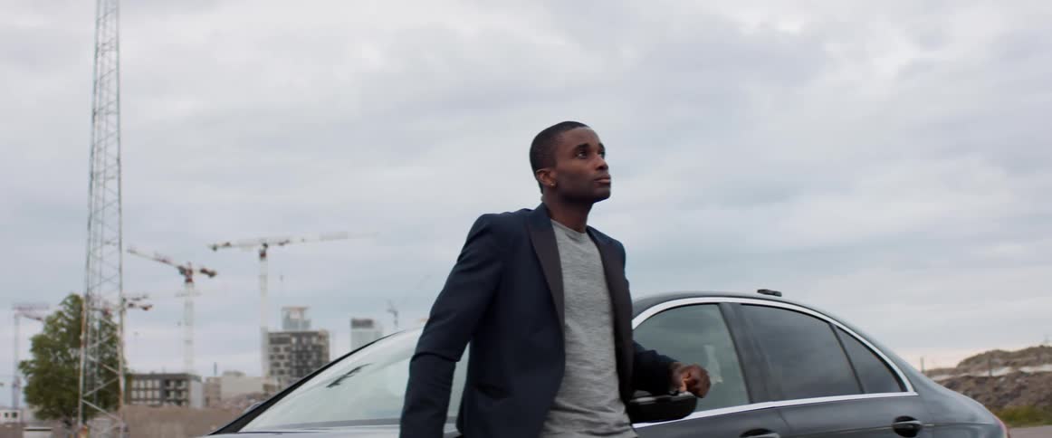 A young man leans against the hood of a car while looking up at the cloudy daytime sky