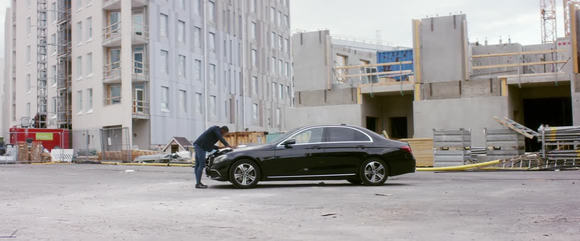 Man in business jacket and jeans leans over black sedan consulting paper near building under construction