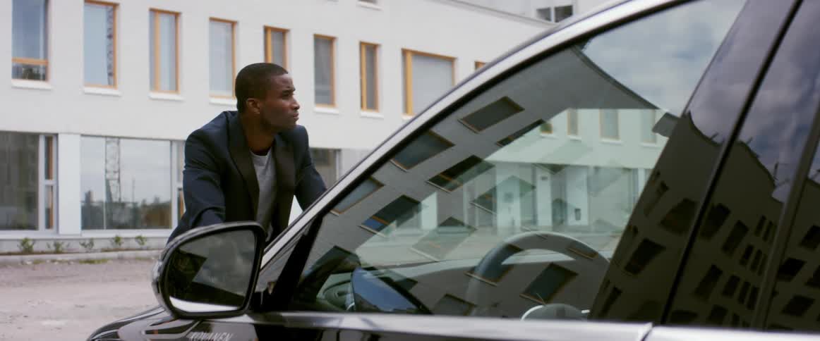A man in front of an apartment building leans on the hood of his car and looks around