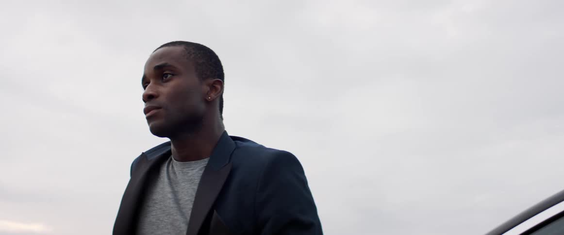 A black man wearing a black dress jacket over a gray shirt is standing and looking ahead under a gray sky