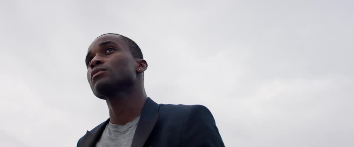 Man who is wearing a grey shirt and blue jacket and an earring is standing still in the wind and looking into the distance with a grey sky behind him