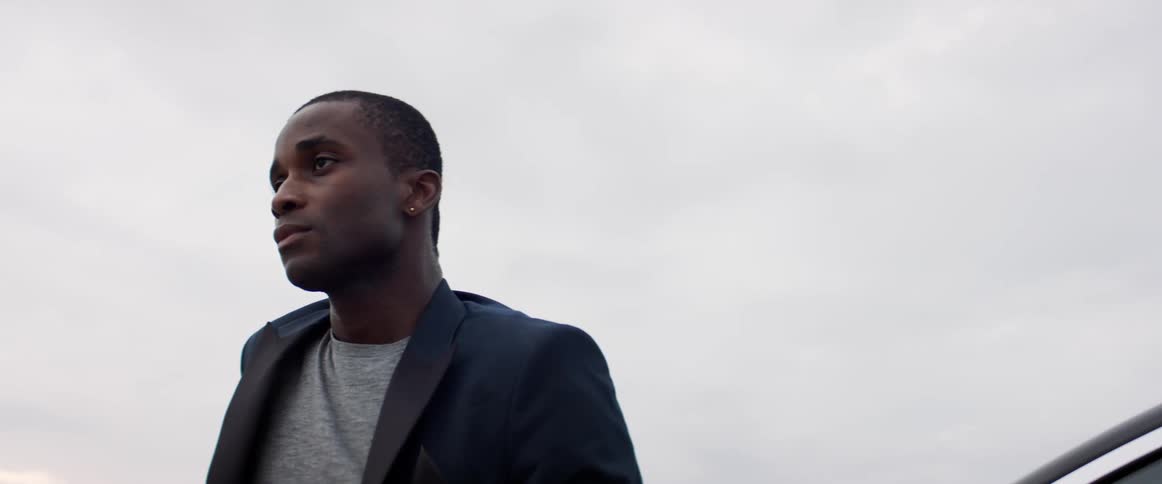 A young black man standing outside on a cloudy day with his eyes closes opens them and stares off in to the distance