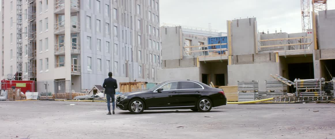 A man gets out of his car at a construction site and walks to the front of his car and spreads blueprints on the hood
