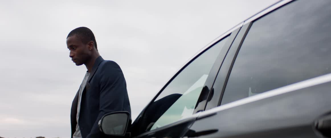 A black man wearing a blazer leans against a black car outside on an overcast day