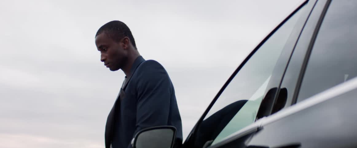 A man stands against a vehicle with a somber expression on his face while lost in thought
