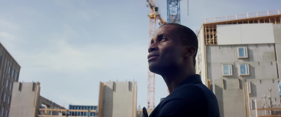Man in business suit stands looking up at buildings near building under construction by crane