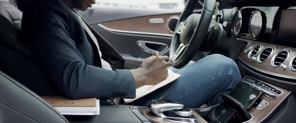 Man wearing suit blazer and jeans sits in drivers seat writing in notebook