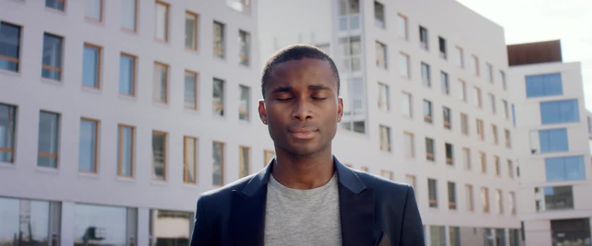 A man standing outdoors in a metropolis during the day stares emotionlessly into the camera