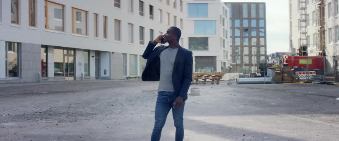 A man standing outdoors on a construction site in a city gestures at the buildings around him while talking on his smartphone