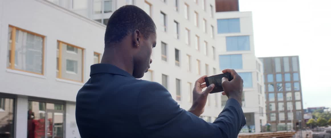 A man standing in a city holding a smart phone with buildings the sky and clouds in the background