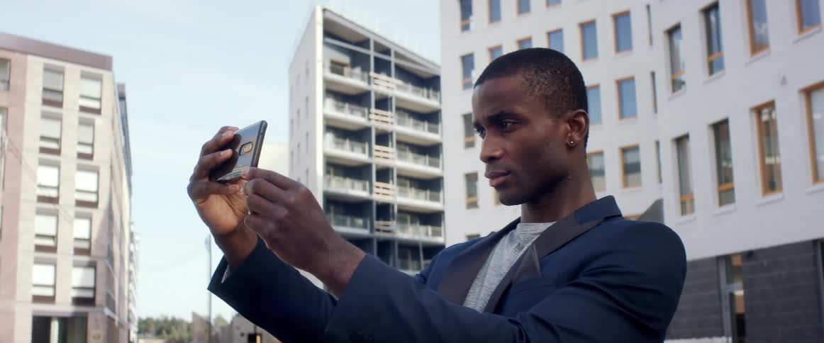 A man standing in a city taking a picture with a smart phone with buildings the sky and clouds in the background