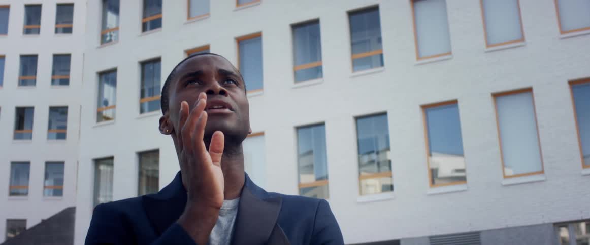 A young black man stands outside apartment buildings and looks at them