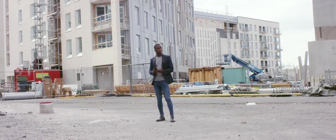 A man stands at a construction site in the city and looks at his phone and then looks at the buildings around him