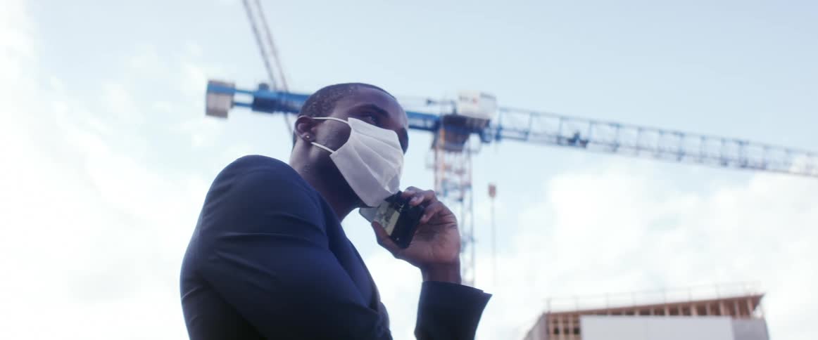 Man wearing business suit and face mask talks on phone near building under construction by crane