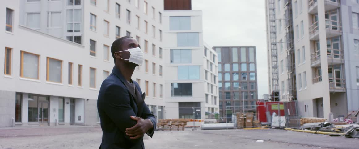 A man stands outside of large city buildings wearing a face mask