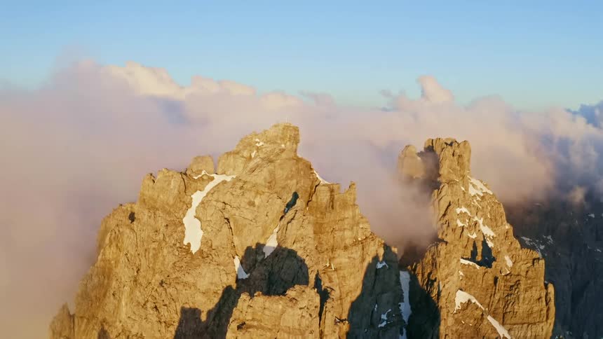 Pink tinted clouds hover over stone mountain top dotted with snow