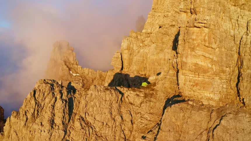 Aerial view of a person standing next to their tent on the edge of a cliff on a mountain range