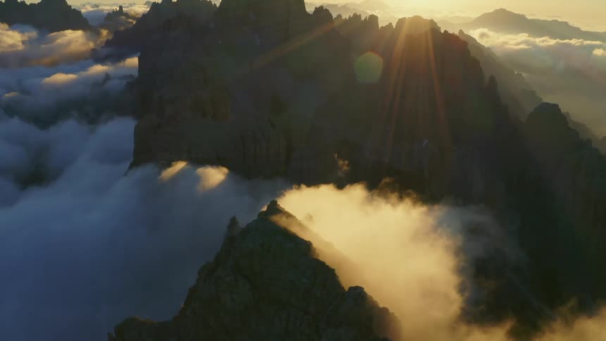 A mountain range peaks are surrounded with clouds and the rays of the rising orange sun
