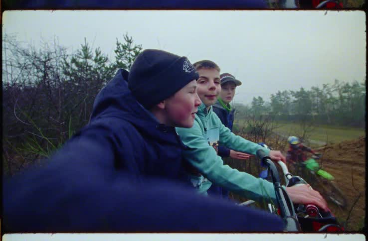 Three young boys on bikes yell and cheer on people racing on motorcycles