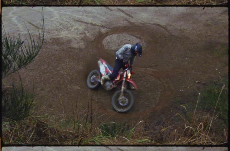A motorcyclist does donuts in the soil on a dirt bike before riding out of frame
