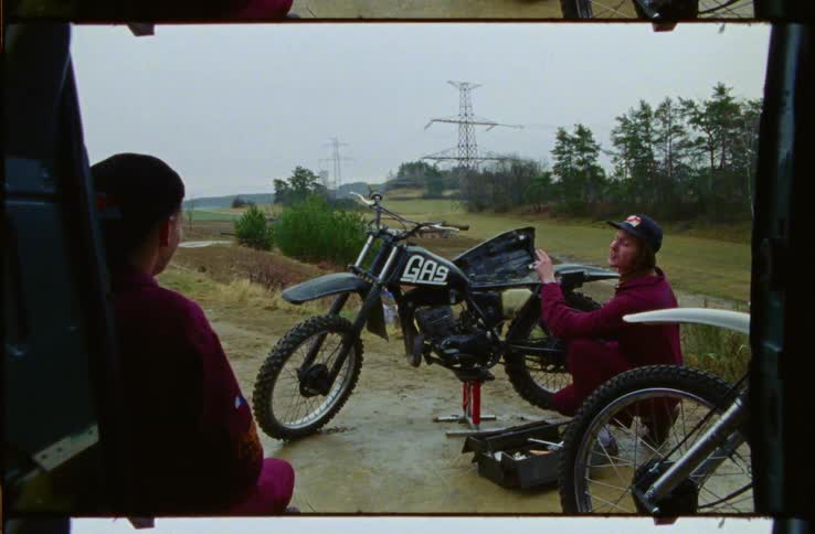 Two mechanics relax near motorcycles and toolbox as motorcyclists race along course on foggy day