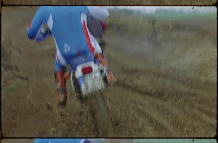 Motocross riders ride up dirt mound past leafless trees as mist obscures horizon