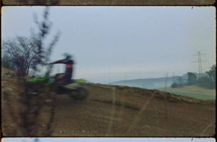Motocross competitors race along sloping dirt track as fog sits near hills on horizon