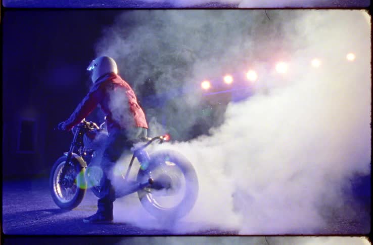 Person in a helmet red coat jeans and boots sits on a motorcycle outside of some buildings in a lit up parking lot on a starry night burning tires