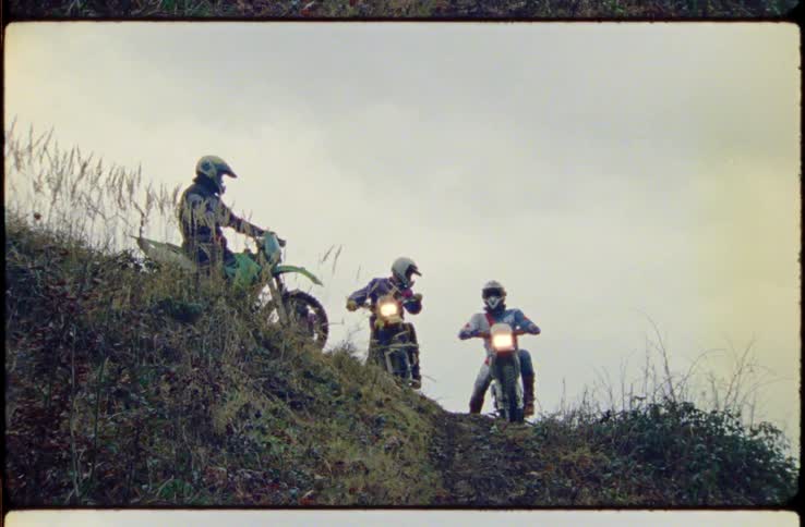People with helmets riding dirt bikes on a hill with the sky and clouds in the background