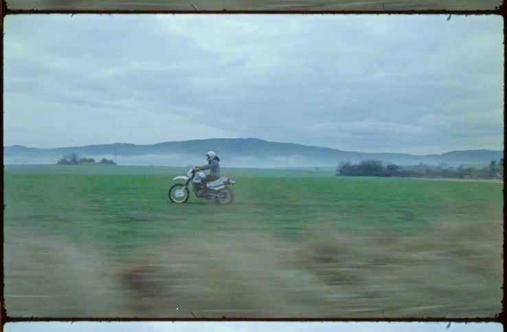 A motorcyclist rides a dirt bike off road through a field of grass on a cloudy day