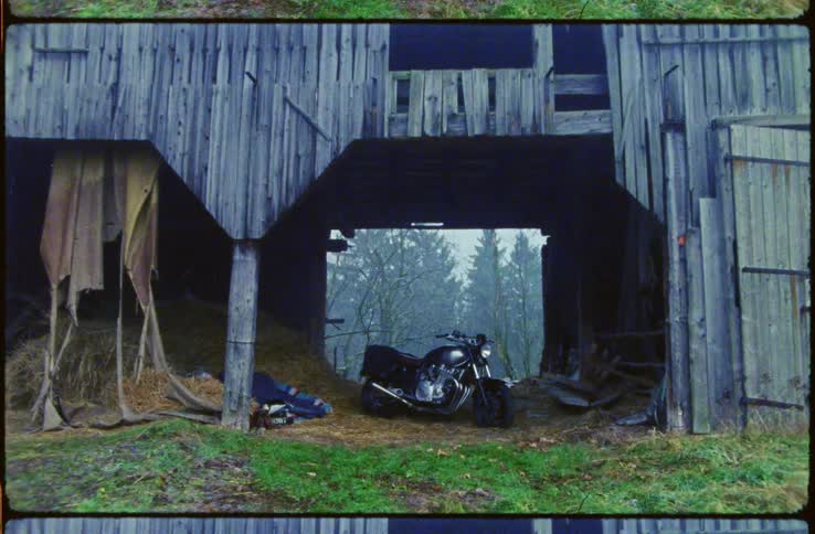 Man in a sleeping bag on hay sleeping next to a black motorcycle inside an old wooden barn