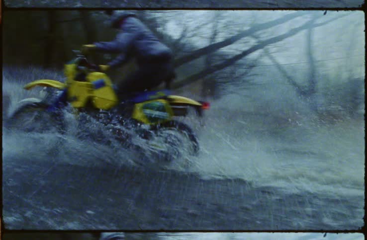 Two motorcyclists race along stream bed past leafless trees and hills
