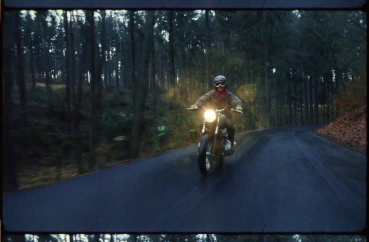 Person rides motorcycle along winding road past woods and hills