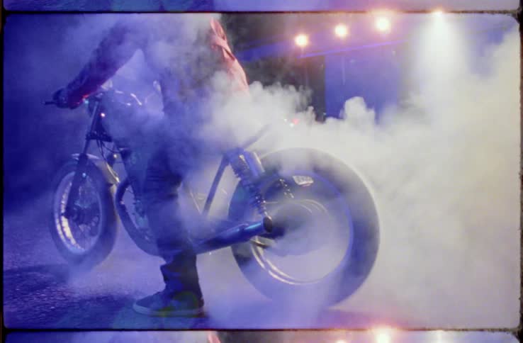 Person sits motorcycle with one foot on ground keeping it in place burning rubber creating smoke under lights