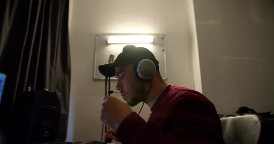 A man with headphones and a hat sitting at a desk making music on a computer