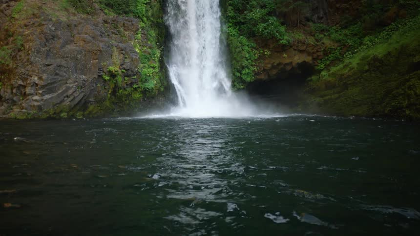 Waterfall splashes into rippling water reflecting the falls down steep vegetation covered slopes